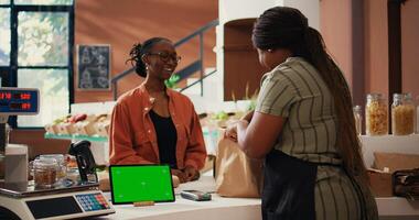 Customer approaching counter with greenscreen on device, buying eco friendly goods from local owner. African american vendor using isolated copyspace display on tablet, talks to client. Tripod shot. photo