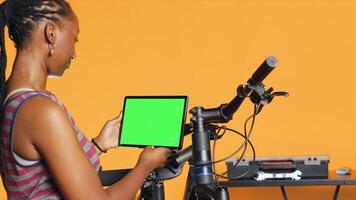 Joyful woman following tutorial videos on mockup tablet, learning to mend broken bike. Smiling cycling hobbyist having fun repairing bicycle, following online guide on green screen device, camera A photo