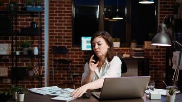 Woman answers phone call from husband while working in office at computer desk. Employee using smartphone to chat with friend during telephone call while at work in front of laptop at night, camera A photo