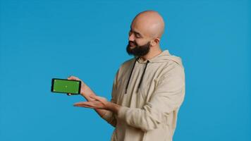 Middle eastern man presenting greenscreen on smartphone, showing chromakey display with isolated mockup layout in studio. Muslim adult creating promotion with blank copyspace screen. Camera 1. photo
