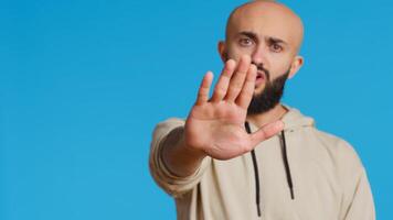 Middle eastern man raises palm and says no in studio, expressing his rejection with stop symbol over blue background. Negative person showing denial and restriction. Camera 2. Handheld shot. photo