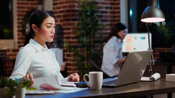 Asian bookkeeper working in office overnight, bored of checking financial data graphs and charts. Businesswoman trying to finish tedious paperwork, feeling stressed and extenuated photo