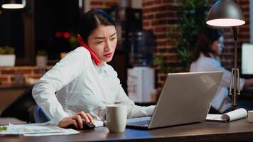Company secretary taking phone call at office desk, answering customer inquiries while working on laptop, multitasking. Stressed worker having conversation on telephone and crosschecking data photo