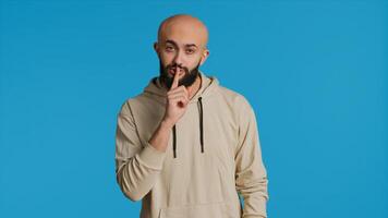 Middle eastern man doing hush silence symbol in studio, asking to keep a secret over blue background. Arab person creating mute gesture to be quiet in front of camera. Camera 1. photo