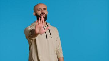 Middle eastern man raises palm and says no in studio, expressing his rejection with stop symbol over blue background. Negative person showing denial and restriction. Camera 1. photo