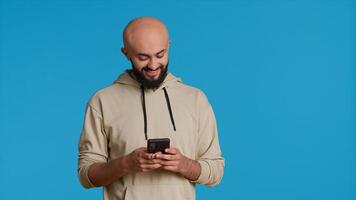 Arab person typing text messages on smartphone app, using mobile phone internet to check social media. Middle eastern guy browsing online websites in studio, blue backdrop. Camera 1. photo