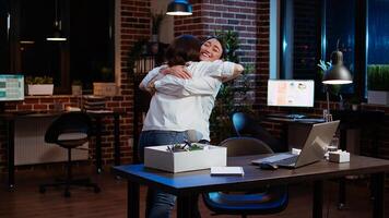 Woman packing office desk belongings, excited to go on holiday, hugging asian coworker. Joyful employee putting objects on workspace table in box, taking days off to go on vacation, camera A photo