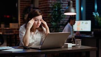 Tired employee struggling to remain focused at workspace desk, feeling overworked and waiting to go home. Bored businesswoman sighing in office while working overnight, camera B photo
