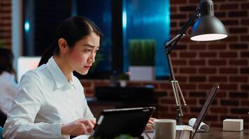 Businesswoman crosschecks data between financial charts and company internet website, doing tasks for team project in office overnight. Employee works at night, typing on laptop keyboard, camera B photo