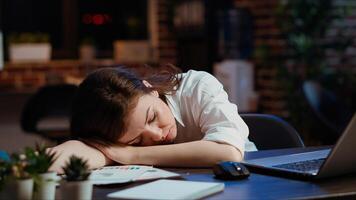 Sleepy employee asleep on computer desk late at night in brick wall office. Panning shot of tired businesswoman sleeping with head on table while working overnight on laptop, camera B photo