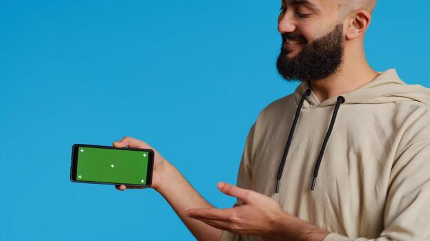 Middle eastern man presenting greenscreen on smartphone, showing chromakey display with isolated mockup layout in studio. Muslim adult creating promotion with blank copyspace screen. Camera 2. photo