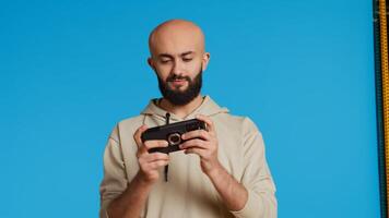 Joyful muslim man playing mobile games in studio, enjoying online competition using smartphone app. Arabic gamer having fun with roleplaying challenge contest. Camera 2. Handheld shot. photo
