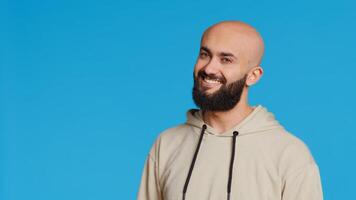 Middle eastern guy greeting people in front of camera, waving hello and smiling over blue background. Young person posing with confidence in studio, feeling confident and joyful. Camera 2. photo