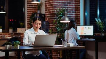 Smiling asian worker researching key data for company project, doing tasks in office overnight. Cheerful staff member looking over accounting figures on laptop screen late at night, typing on keyboard photo