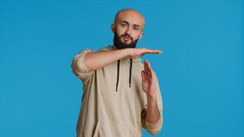 Middle eastern person doing timeout gesture on camera, asking for a break to stop working over blue background. Muslim adult presents symbol to pause or stop something. Camera 1. photo