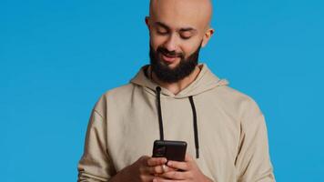 Arab person typing text messages on smartphone app, using mobile phone internet to check social media. Middle eastern guy browsing online websites in studio, blue backdrop. Camera 2. photo