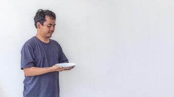 Adult man holding and looking at an empty white plate with a hungry expression, depicting poverty, isolated on a white background. Ideal for themes related to hunger, food scarcity, and social issues. photo