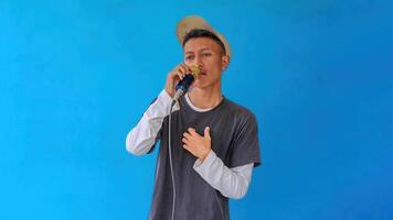 Young expressive singer happy man wearing gray t-shirt with glasses and hat singing song into microphone spreading hands isolated on plain light blue background studio. People's lifestyle concept photo