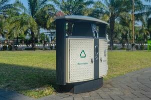 A modern, public recycling bin with a clear label and a green recycling symbol, placed in a park setting with palm trees in the background. photo