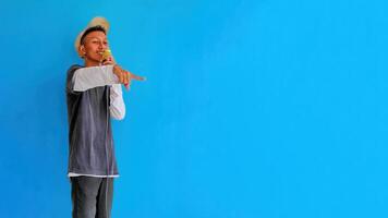 Young expressive singer happy man wearing gray t-shirt with glasses and hat singing song into microphone spreading hands isolated on plain light blue background studio. People's lifestyle concept photo