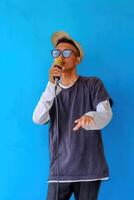 Young expressive singer happy man wearing gray t-shirt with glasses and hat singing song into microphone spreading hands isolated on plain light blue background studio. People's lifestyle concept photo