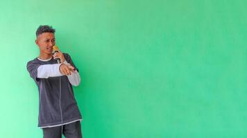 Young expressive singer happy man wearing gray t-shirt with glasses and hat singing song into microphone spreading hands isolated on plain light green background studio. People's lifestyle concept photo