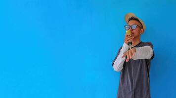 Young expressive singer happy man wearing gray t-shirt with glasses and hat singing song into microphone spreading hands isolated on plain light blue background studio. People's lifestyle concept photo