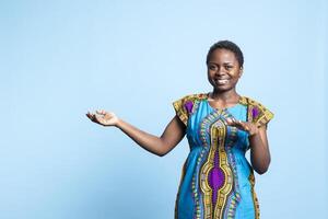 Charming person wears in ethnic attire and shows something in studio, creates advertisement, pointing to the left or right. African american woman does web advertisement on camera. photo