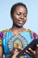 Modern casual person is using a portable device to browse social media pages over a blue background. African american woman searches information on tablet in front of the camera. photo