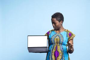 Delighted woman in studio holding white notebook screen with isolated mockup setup on it. Happy girl wearing ethnic attire, holding laptop with a blank copyspace display, web commercial. photo