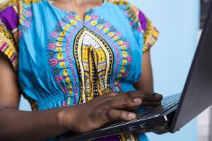 African american model navigating on internet webpages over blue background, using wireless computer. Young adult dressed in ethnic tribal clothing items uses laptop to check social media. photo
