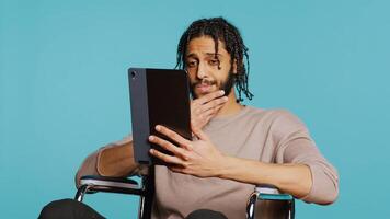 Person with paraplegia using tablet to post images on social media, doing selfie and writing photo caption. Man in wheelchair using online editing application on digital device, studio, camera B