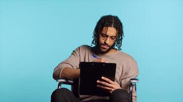 Middle Eastern man in wheelchair suffering from paraplegia holding clipboard in hand, using pen to write on paper. Person with disability completing forms, isolated over studio background, camera A photo