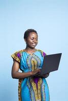 Young online user works on notebook computer to access internet sites while exploring social media in a studio. African american girl smiles for the camera while she is holding electronic device. photo