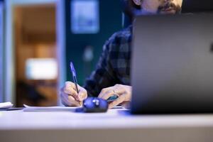 A man works from home, using his laptop and taking notes on a notepad at a modern desk in his apartment. Close-up of a bearded man writing in a notebook while studying and researching. photo