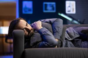 Man laying down hugging pillow watching TV, heartbroken by glum movie plot. Sad viewer lounged on couch in RGB illuminated apartment, upset by depressing television show scene photo