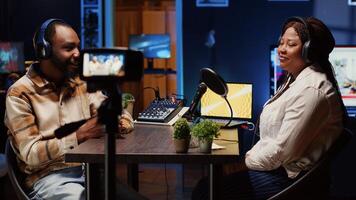 African american influencer using professional camera to record podcast with guest, talking and having fun together. Happy host discussing with woman live in apartment studio for internet show photo