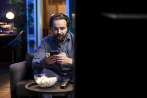 Man enjoying bowl of popcorn while watching cable programs on TV and texting friends on phone. Spectator on couch in living room eats snacks and talks with mates on smartphone photo