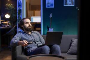 Man watching interesting movie on streaming service with laptop on lap, enjoying time off from work. Person on couch consuming snacks, looking at films broadcasted on TV and holding notebook photo