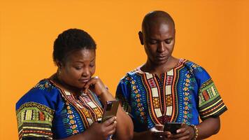 Ethnic couple scrolling on social media accounts in studio, using smartphones to navigate online network webpages. African american people in traditional attire checking internet sites. photo