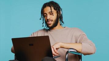 Man in wheelchair chatting with friend over internet videocall using laptop application. BIPOC person with paraplegia chatting with mate in videoconference online meeting, studio background, camera B photo