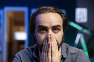 Portrait of cord cutter covering mouth in shock while looking in disbelief at TV displaying thriller film. Disturbed person at home watching movie on on demand channel, stupefied by plot twist photo