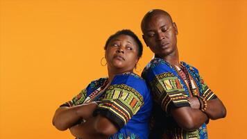 Carefree relaxed ethnic couple posing on camera, standing back to back and wearing traditional colorful clothes. African american people feeling confident and joyful together over orange background. photo
