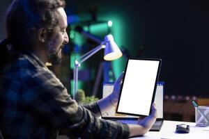 Bearded man sitting at desk, grasping a tablet showing an isolated white screen with laptop in the background. Male freelancer using digital device displays a blank mockup template. photo