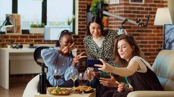 Multiracial friends taking group pictures with smartphone at apartment gathering. Cheerful people in living room posing for photo, making gestures and drinking alcoholic beverages photo