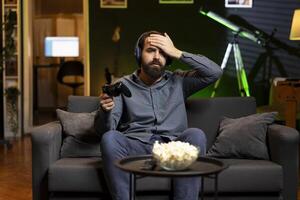 Gamer wearing headphones face palming himself in frustration after getting game over message while playing videogame. Man feeling depressed while playing on gaming console after losing photo