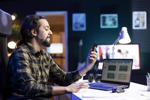 Bearded guy holding and using his smartphone, checking his social media and answering messages. Male individual sitting at table with laptop while looking at his mobile device. photo