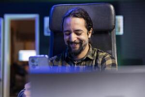 Male freelancer taking a break, cheerfully using his smartphone to watch funny online content. Positive young man sitting at desk laughing while texting on his mobile device, surfing the net. photo