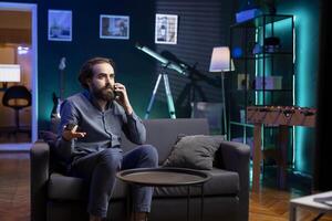 Sad supporter watching in disbelief own team getting defeated during match broadcast, discussing with friend on phone. Man heartbroken by favorite club failing to win, talking with mate on smartphone photo
