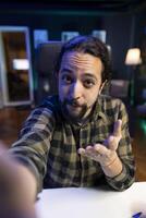 Male individual gesturing towards the camera, having a live online conversation with his social media groups. POV of person holding a smart gadget, recording a at home. photo